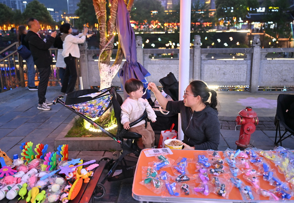 4月12日，在宣恩縣城貢水河畔，一名擺地?cái)偟膵D女在給小孩喂飯。